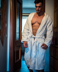 Full body muscular guy in untied white bathrobe looking at camera while standing in hallway in...