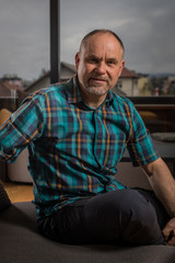 Fototapeta premium A caucasian man in his late forties with short hair and short gray beard is sitting in a couch and looking into the camera. Forest is seen behind the window.