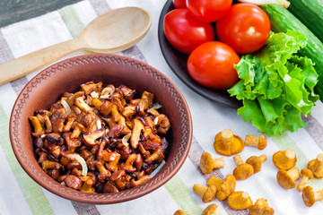 Fried chanterelles with onion in rustic bowl and plate with fresh vegetables for salad on background