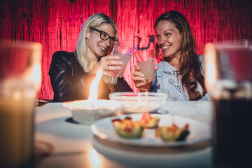 Two young women, a blonde and a redhead are cheering with glasses full of gin tonic drink and straws on a garden pary with candle on early evening - obrazy, fototapety, plakaty