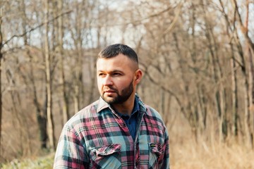 Bearded hipster man with serious face is thinking about something while standing outdoors against autumn fall with copy space, stylish brutal male with trendy look walking in park at sunset.