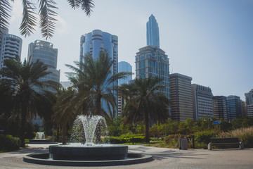 Fototapeta na wymiar Cityscape of Abu Dhabi on a clear cunny day with a fountain and some palms in the foregroud and with majestic glass skyscrapers in the background.