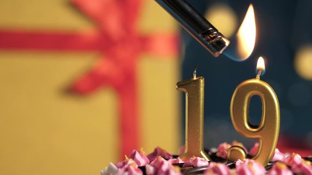 Birthday cake number 19 golden candles burning by lighter, blue background gift yellow box tied up with red ribbon. Close-up and slow motion