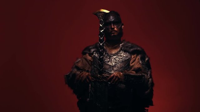 A warrior with a bloody head throws an axe on his shoulder, looking at the camera from under his brows. The bearded Viking puts his battle axe on his shoulder, looks aggressively at the camera