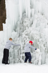Two little child play under frozen waterfall, exploring the strange ice formations.