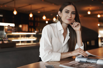 Elegant young businesswoman reading magazine while sitting in cafe, leaning desk, wearing white...