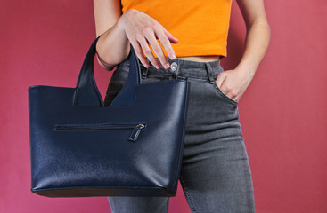 Fashionable leather bag hanging on woman's hand with orange T-shirt and gray jeans on red background. Crop photo, studio shot