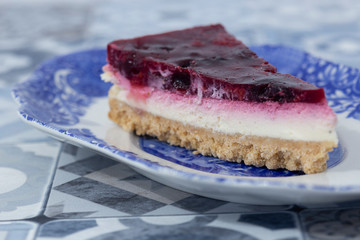 Slice of summer fruit cheesecake with a biscuit base on a blue and white ornate plate. With a blue patterned tiled worktop background
