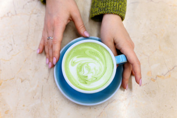 matcha green tea latte with a pattern of milk foam in a blue ceramic cup and women's hands on the table