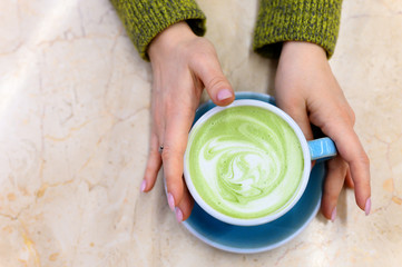 matcha green tea latte with a pattern of milk foam in a blue ceramic cup and women's hands on the...
