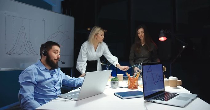 Caucasian handsome man in headphones and mic having videochat at laptop computer in call center. Male worker of call-center speaking with client in evening. Females co-workers talking behind.