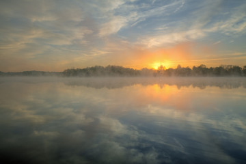 Fototapeta na wymiar Landscape at sunrise of Whitford Lake in fog with mirrored reflections in calm water, Fort Custer State Park, Michigan, USA
