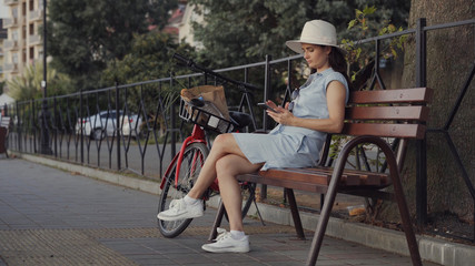 A woman in a hat and dress sits on a bench near a red bicycle and uses her phone.
