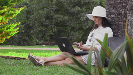 A woman in a hat sits on the grass in a Park with a laptop and uses a phone.