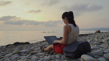 A woman at sunset behind a laptop works on the beach.
