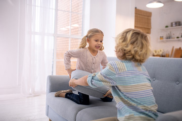 Two blond kids playing with grey sofa pillows