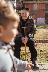 Senior old man caucasian pensioner grandfather holding his cane on the sitting on the bench by sidewalk pavement in park sunny winter or spring day while boy child kid is running in front of him