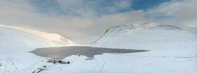 An aerial panoramic scenic view of a mountain lake in the winter with snowy slope under a majestic blue sky and white clouds