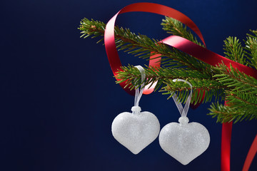 Still life for Valentine's day two silver hearts on a coniferous branch with a red ribbon on a dark blue background, space for text.