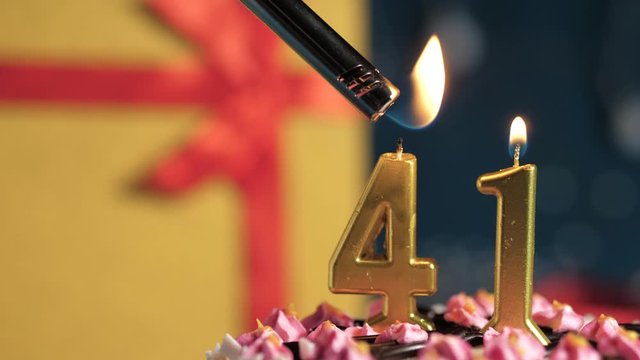 Birthday cake number 41 golden candles burning by lighter, blue background gift yellow box tied up with red ribbon. Close-up and slow motion