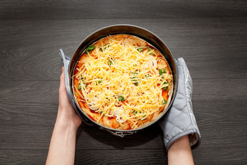 The pizza is strewn with a large amount of cheese, in the hands of a potholder, on a wooden background, before being sent to the oven. The concept of cooking pizza.