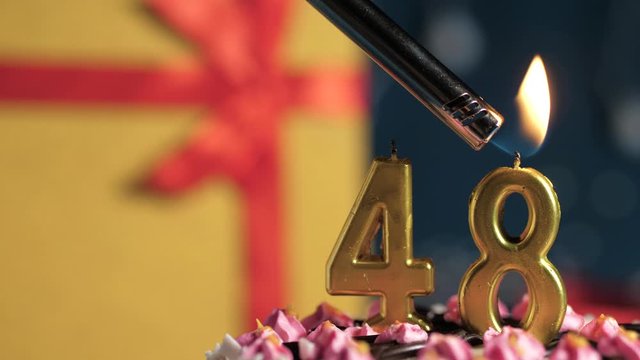 Birthday cake number 48 golden candles burning by lighter, blue background gift yellow box tied up with red ribbon. Close-up and slow motion