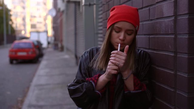 Young Beautiful stylish European girl 30s years dressing black leather jacket jeans and red hat standing on backyard near building and smoking cigarette Stress concept Bad habit unhealthy lifestyle.