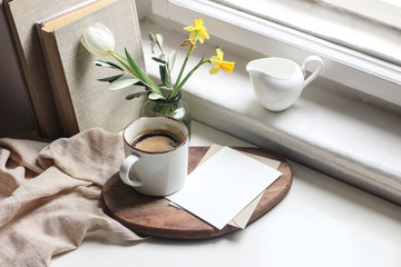 Cozy Easter spring still life. Greeting card mockup scene. Cup of coffee, books, wooden cutting board, milk pitcher and vase of flowers on windowsill. Floral composition. Yellow daffodils and tulip.