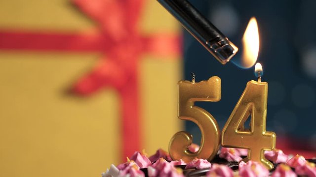 Birthday cake number 54 golden candles burning by lighter, blue background gift yellow box tied up with red ribbon. Close-up and slow motion