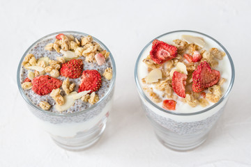 Two glasses of overnight chia seed pudding with almond milk, yogurt, homemade granola, dried strawberries and almonds. Superfood and vegan food concept. Copy space, selective focus, white background