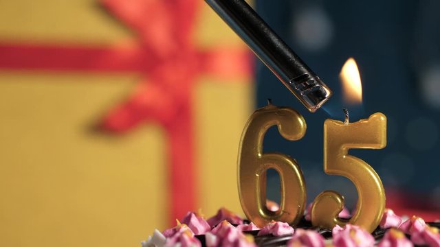 Birthday cake number 65 golden candles burning by lighter, blue background gift yellow box tied up with red ribbon. Close-up and slow motion