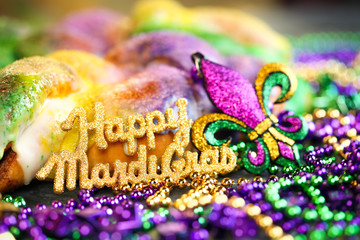 Happy Mardi Gras text in gold glitter and a king cake with yellow, green, and purple sprinkles surrounded by Mardi Gras beads and a glittering fleur de lis.