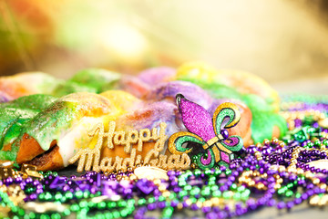 Fototapeta Happy Mardi Gras text in gold glitter and a king cake with yellow, green, and purple sprinkles surrounded by Mardi Gras beads and a glittering fleur de lis. obraz