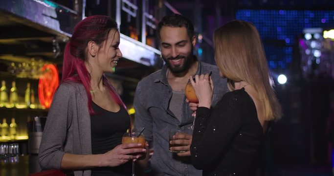 Middle Eastern man coming to two young Caucasian women drinking cocktails in night club. Confident handsome guy flirting with girls in disco. Cinema 4k ProRes HQ.