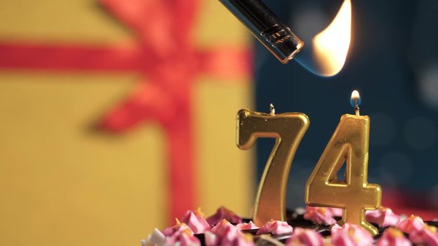 Birthday cake number 74 golden candles burning by lighter, blue background gift yellow box tied up with red ribbon. Close-up and slow motion