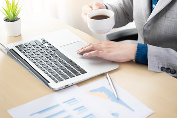 Businessman is drinking coffee in the morning while working on financial reports using his laptop computer. Relaxing time while working concept.