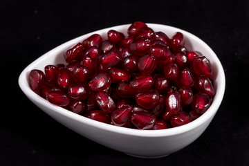 Refined pomegranate: juicy shiny pomegranate seeds are poured into a small bowl and photographed against a black background in the studio. The combination of three colors: red, black and white. Backgr