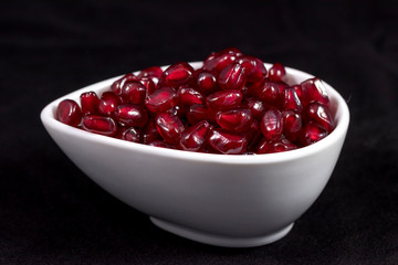 Refined pomegranate: juicy shiny pomegranate seeds are poured into a small bowl and photographed against a black background in the studio. The combination of three colors: red, black and white. 