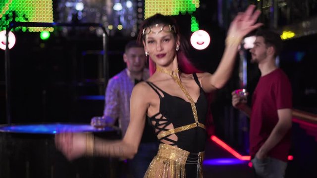 Portrait of young confident Caucasian woman dancing and spinning as blurred men standing at the background. Beautiful woman resting in night club. Joy, lifestyle, leisure.