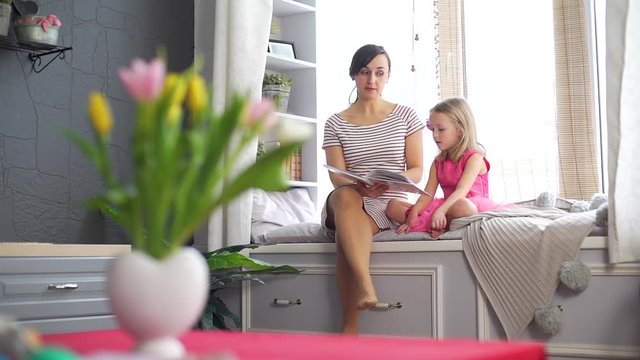 Young Mother Reading Book to her Cute Little Daughter at Home. They Sitting on the Big Cozy Windowsill. Slow Motion. People, Lifestyle and Happy Family Concept