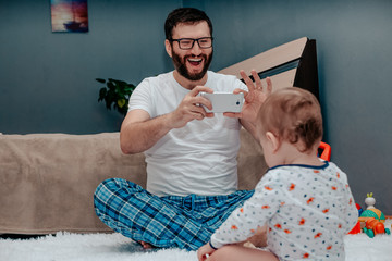 Father in home clothes sitting on the floor photographs the child on the phone. The parent is very...