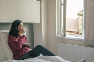 Woman is drinking hot morning coffee on her bed int the apartment.