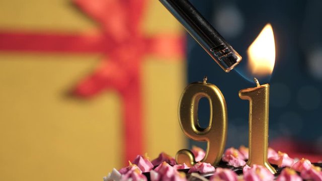 Birthday cake number 91 golden candles burning by lighter, blue background gift yellow box tied up with red ribbon. Close-up and slow motion
