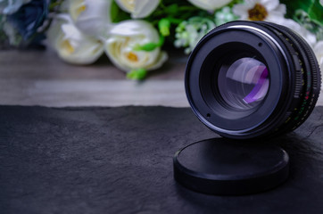 An old lens for a camera with a focal length of 50 mm stands on a table and is turned towards the camera with flowers in the background. For advertising the photographer.