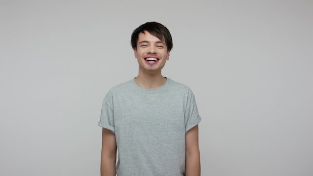 What a funny story! Young brunette guy in T-shirt laughing out loud and wiping tears from hilarious laughter, feeling amused after hearing anecdote, ridiculous joke. indoor studio shot gray background