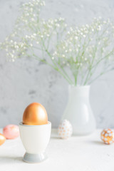 Close up of Golden egg in egg cup on the background of bouquet of gypsophila. Cozy easter still life