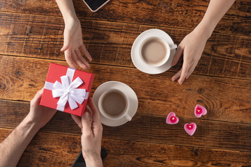 Valentine's Day celebration concept. A nice gift for your loved one. Hands of man and woman with coffee mugs on a wooden table background. Copy space. Flat lay. Close-up.
