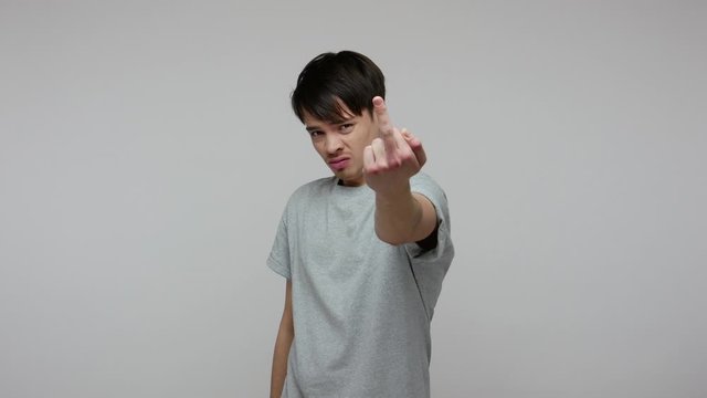 Crazy rude vulgar man in T-shirt showing middle fingers with both hands and saying fuck you, expressing protest aggression hate, impolite behavior. indoor studio shot isolated on gray background