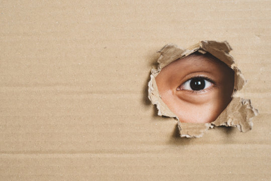 Boy eye peeking through a hole on a cardboard box with shocking gesture. Concept of mystery, secret and stalker