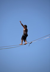 Akrobata spacerujący po linie zawieszonej na dużej wysokości na tle niebieskiego niba - obrazy, fototapety, plakaty